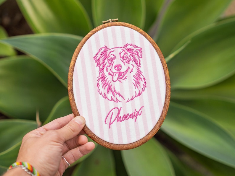 May include: An oval wooden embroidery hoop containing white fabric with vertical light pink stripes. The fabric features a magenta embroidered portrait of a dog with its tongue out, positioned above the cursive magenta text reading Phoenix.