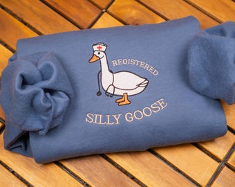 May include: A slate blue sweatshirt featuring an embroidered graphic of a white goose wearing a nurse cap and a stethoscope. The text above the goose reads REGISTERED and the text below reads SILLY GOOSE in light-colored, embroidered lettering.