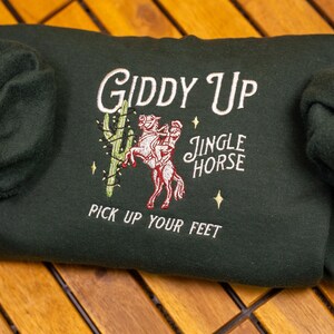 May include: A dark green sweatshirt with an embroidered design of a person riding a horse with the text "Giddy Up Jingle Horse Pick Up Your Feet".
