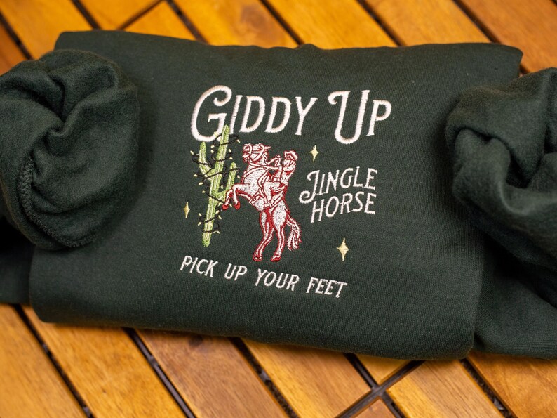 May include: A dark green sweatshirt with an embroidered design of a person riding a horse with the text "Giddy Up Jingle Horse Pick Up Your Feet".
