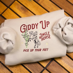 May include: A beige sweatshirt with the text "Giddy Up Jingle Horse Pick Up Your Feet" embroidered in red on the front.