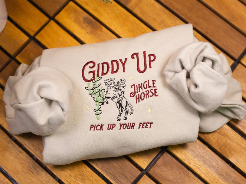 May include: A beige sweatshirt with the text "Giddy Up Jingle Horse Pick Up Your Feet" embroidered in red on the front.