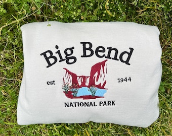 Puede incluir: Una sudadera beige con un diseño bordado del Parque Nacional Big Bend. El diseño incluye el nombre del parque, una imagen estilizada del paisaje del parque y los años "est 1944".