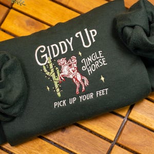May include: A dark green sweatshirt with the text "Giddy Up" and "Jingle Horse" embroidered on the front. The sweatshirt also features an embroidered image of a person riding a horse with a cactus in the background. The text "Pick Up Your Feet" is embroidered below the image.