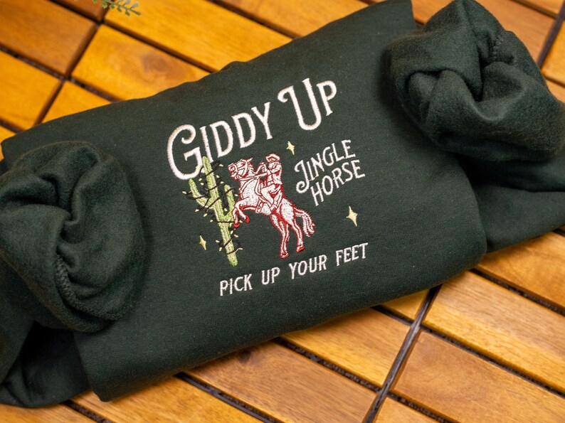May include: A dark green sweatshirt with the text "Giddy Up" and "Jingle Horse" embroidered on the front. The sweatshirt also features an embroidered image of a person riding a horse with a cactus in the background. The text "Pick Up Your Feet" is embroidered below the image.