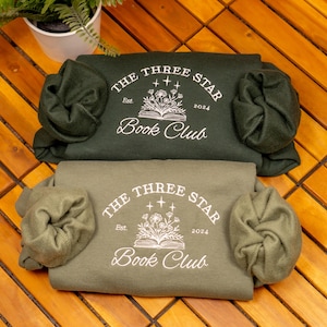 May include: Two folded crewneck sweatshirts in dark forest green and muted olive green. Each features a white screen-printed graphic of an open book with flowers and stars, with the text "THE THREE STAR BOOK CLUB" and "Est. 2024" printed in white.