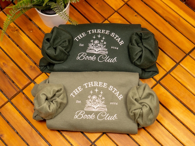 May include: Two folded crewneck sweatshirts in dark forest green and muted olive green. Each features a white screen-printed graphic of an open book with flowers and stars, with the text "THE THREE STAR BOOK CLUB" and "Est. 2024" printed in white.