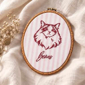 May include: An oval embroidery hoop containing fabric with vertical light pink and white stripes. The fabric features a dark red embroidered portrait of a fluffy cat and the word Beans in cursive script. The hoop has a dark brown wood grain finish.