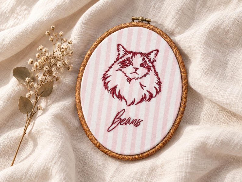 May include: An oval embroidery hoop containing fabric with vertical light pink and white stripes. The fabric features a dark red embroidered portrait of a fluffy cat and the word Beans in cursive script. The hoop has a dark brown wood grain finish.