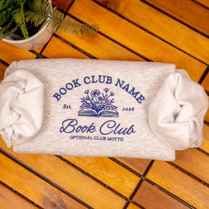 May include: A light gray heather sweatshirt featuring a blue graphic of an open book with flowers emerging from the pages. The text reads BOOK CLUB NAME, Est. 2026, Book Club, and OPTIONAL CLUB MOTTO. The sleeves are gathered at the sides.