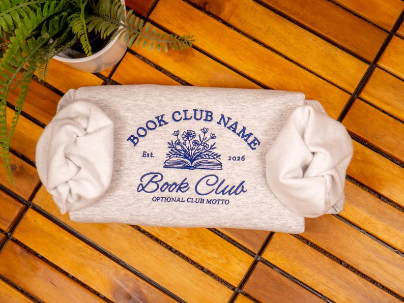 May include: A light gray heather sweatshirt featuring a blue graphic of an open book with flowers emerging from the pages. The text reads BOOK CLUB NAME, Est. 2026, Book Club, and OPTIONAL CLUB MOTTO. The sleeves are gathered at the sides.