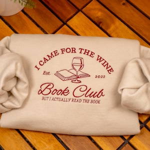 May include: A cream-colored sweatshirt featuring dark red embroidered text that reads I CAME FOR THE WINE, Est. 2022, Book Club, and BUT I ACTUALLY READ THE BOOK. The design includes a central graphic of an open book with a wine glass on top.