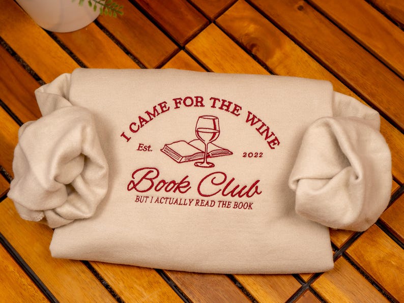 May include: A cream-colored sweatshirt featuring dark red embroidered text that reads I CAME FOR THE WINE, Est. 2022, Book Club, and BUT I ACTUALLY READ THE BOOK. The design includes a central graphic of an open book with a wine glass on top.