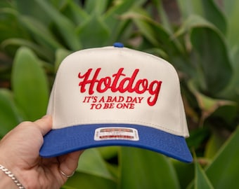 May include: A cream-colored baseball cap with a blue brim and a blue button on top. The word "Hotdog" is embroidered in red script, with the phrase "IT'S A BAD DAY TO BE ONE" below it.