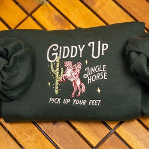 May include: A dark green hooded sweatshirt with the text "Giddy Up Jingle Horse Pick Up Your Feet" embroidered on the front. The design features a cartoon cowboy riding a horse.