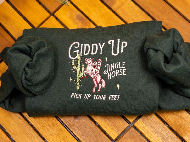 May include: A dark green hooded sweatshirt with the text "Giddy Up Jingle Horse Pick Up Your Feet" embroidered on the front. The design features a cartoon cowboy riding a horse.