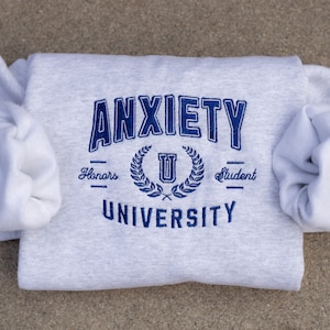 May include: Gray sweatshirt with blue embroidered text that reads "Anxiety University" with a laurel wreath and a "U" in the center. The text "Honors" and "Student" are below the wreath.