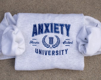 Puede incluir: Sudadera gris con texto bordado en azul que dice "Anxiety University" con una corona de laurel y una "U" en el centro. El texto "Honors" y "Student" está debajo de la corona.