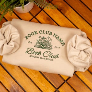 May include: A beige sweatshirt featuring dark green embroidery with the text BOOK CLUB NAME, Est. 2026, Book Club, and OPTIONAL CLUB MOTTO, centered around an illustration of an open book with flowers. Two beige scrunchies are placed on the sides.