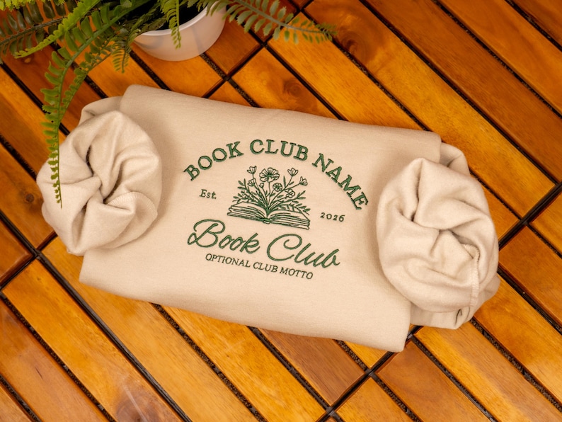 May include: A beige sweatshirt featuring dark green embroidery with the text BOOK CLUB NAME, Est. 2026, Book Club, and OPTIONAL CLUB MOTTO, centered around an illustration of an open book with flowers. Two beige scrunchies are placed on the sides.