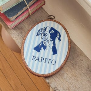 May include: An oval embroidery hoop frame containing fabric with a light blue and white vertical striped pattern. The fabric features a dark blue embroidered portrait of a dog and the text PAPITO in dark blue capital letters below the dog.