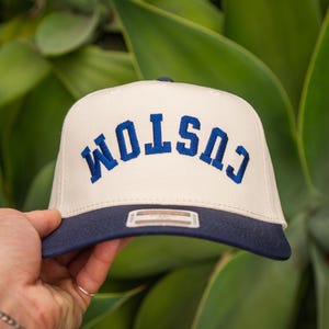 Könnte beinhalten: Eine cremefarbene Baseballkappe mit marineblauem Schirm. Das Wort "CUSTOM" ist in marineblauen Blockbuchstaben auf die Vorderseite der Kappe gestickt. Die Kappe wird vor einem Hintergrund aus grünem Laub gehalten.