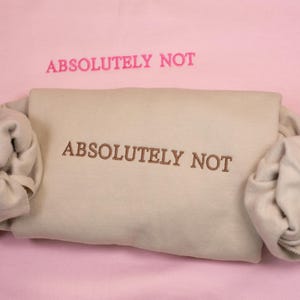 May include: A beige sweatshirt with the phrase "ABSOLUTELY NOT" embroidered in brown on the front. The sweatshirt is folded and displayed on a pink background. The phrase "ABSOLUTELY NOT" is also written in pink on the background.