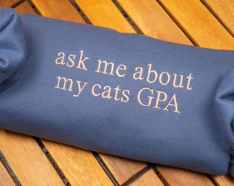 Può includere: Una felpa blu piegata con la scritta ricamata "ask me about my cats GPA" in color beige chiaro. La felpa è appoggiata su una superficie di legno.