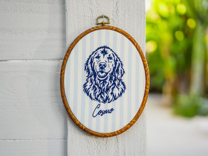 May include: An oval embroidery hoop frame with a wood grain finish containing fabric with light blue and white vertical stripes. A dark blue embroidered portrait of a dog is centered on the fabric with the name Cosmo stitched below in script.