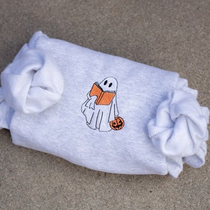 May include: A gray sweatshirt with a white ghost holding a book and a pumpkin. The ghost is embroidered on the sweatshirt.