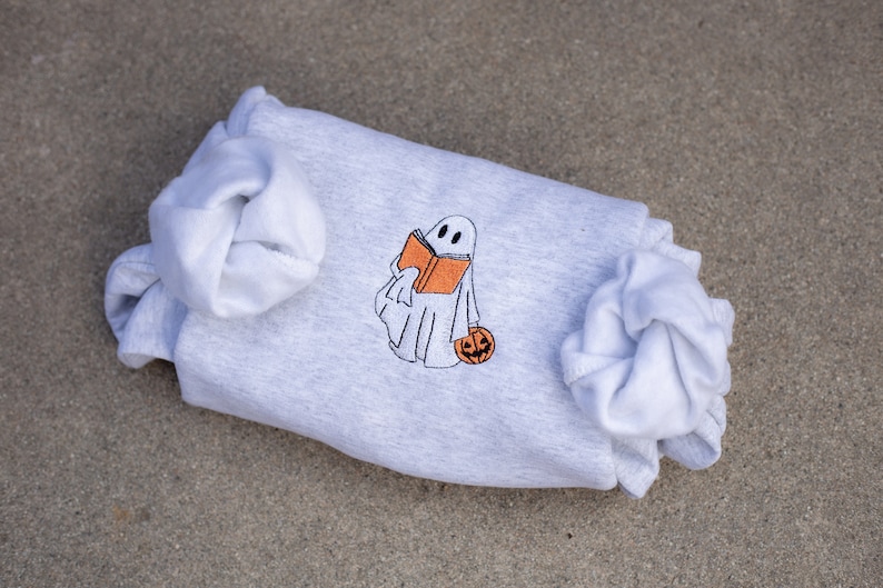 May include: A gray sweatshirt with a white ghost holding a book and a pumpkin. The ghost is embroidered on the sweatshirt.