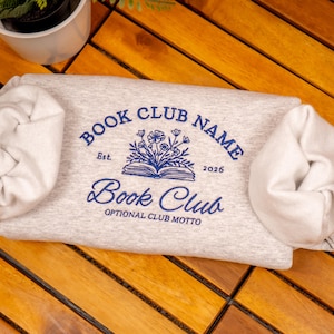 Puede incluir: Una sudadera gris claro con mangas enrolladas. Presenta texto bordado en azul que dice "BOOK CLUB NAME" encima de un diseño floral y de libro, y "Book Club" debajo. También incluye el año "2026".