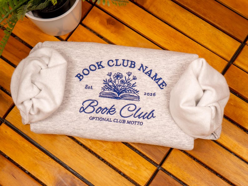 May include: A light gray sweatshirt featuring a blue graphic print. The design includes the text "BOOK CLUB NAME", "Est. 2026", "Book Club", and "OPTIONAL CLUB MOTTO" surrounding an illustration of an open book with flowers emerging from the pages.