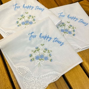 May include: Three white handkerchiefs with embroidered floral bouquets and the text "For happy tears" in light blue. One handkerchief features a butterfly lace edge. The handkerchiefs are arranged on a wooden surface.
