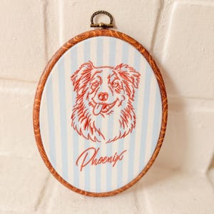 May include: An oval embroidery hoop featuring a light blue and white striped fabric background. Centered is a red stitched outline of a dog's face with its tongue out. Below the dog, the name Phoenix is embroidered in red cursive script.