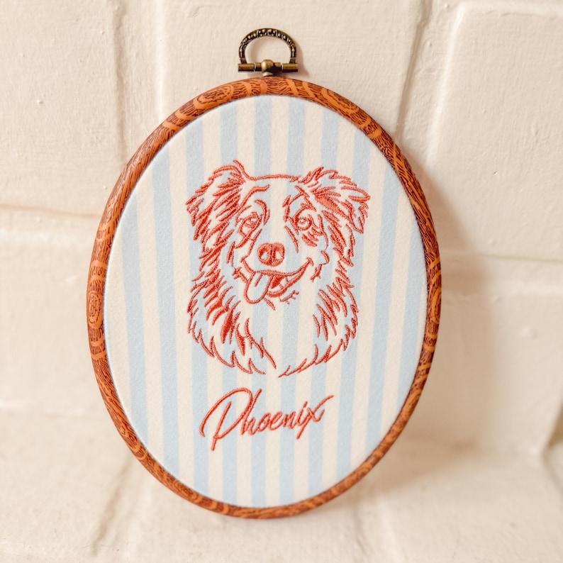 May include: An oval embroidery hoop featuring a light blue and white striped fabric background. Centered is a red stitched outline of a dog's face with its tongue out. Below the dog, the name Phoenix is embroidered in red cursive script.