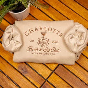 May include: A beige sweatshirt featuring brown text and a graphic of a martini glass resting on a stack of books. The text reads CHARLOTTE, Est. 2026, Book & Sip Club, and MAIN CHARACTERS ONLY. The sleeves are rolled inward toward the center.