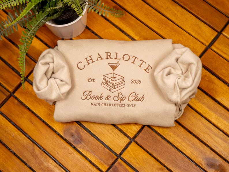 May include: A beige sweatshirt featuring brown text and a graphic of a martini glass resting on a stack of books. The text reads CHARLOTTE, Est. 2026, Book & Sip Club, and MAIN CHARACTERS ONLY. The sleeves are rolled inward toward the center.