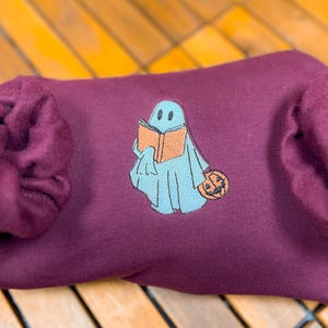 May include: A maroon sweatshirt with a blue ghost holding a book and a pumpkin. The ghost has a friendly face and is wearing a blue and orange book.