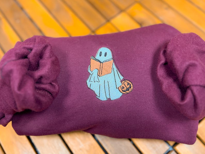 May include: A maroon sweatshirt with a blue ghost holding a book and a pumpkin. The ghost has a friendly face and is wearing a blue and orange book.