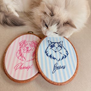 May include: Two oval embroidery hoops with wood-look frames. One features pink vertical stripes with a pink embroidered dog portrait and the name Phoenix. The other has blue vertical stripes with a blue embroidered cat portrait and the name Beans.