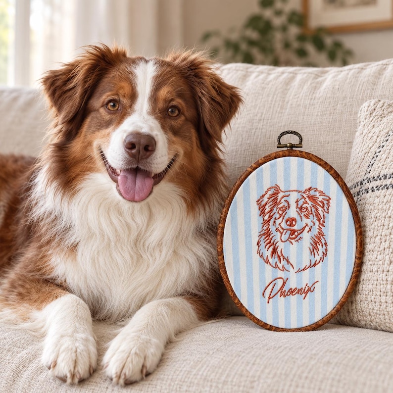 May include: An oval embroidery hoop featuring a light blue and white striped fabric background. Centered on the fabric is a red stitched outline of a dog's face with the name Phoenix embroidered in red cursive script below the dog illustration.