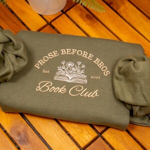 May include: An olive green sweatshirt featuring a graphic print in light gold ink. The design includes an open book with flowers emerging from the pages, surrounded by the text "PROSE BEFORE BROS", "Est.", "2025", and "Book Club".