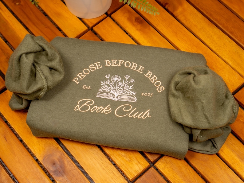 May include: An olive green sweatshirt featuring a graphic print in light gold ink. The design includes an open book with flowers emerging from the pages, surrounded by the text "PROSE BEFORE BROS", "Est.", "2025", and "Book Club".