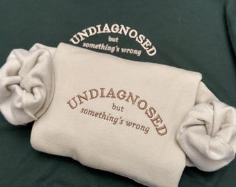 May include: A cream-colored sweatshirt with the text "UNDIAGNOSED but something's wrong" embroidered in brown. The sweatshirt is folded in half and has a green background.