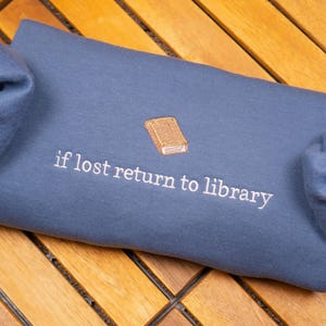 May include: A blue sweatshirt with the text "if lost return to library" and a small embroidered image of a book.