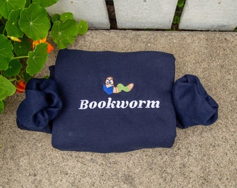 Puede incluir: Sudadera azul marino con la palabra "Bookworm" en blanco. Encima del texto, un gusano de dibujos animados bordado con gafas y un libro. La sudadera está doblada sobre una superficie de hormigón.