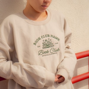 May include: A beige crewneck sweatshirt featuring dark green embroidery on the chest. The design includes an open book with flowers, the text "BOOK CLUB NAME", "Est. 2026", "Book Club", and "OPTIONAL CLUB MOTTO" in a mix of serif and script fonts.