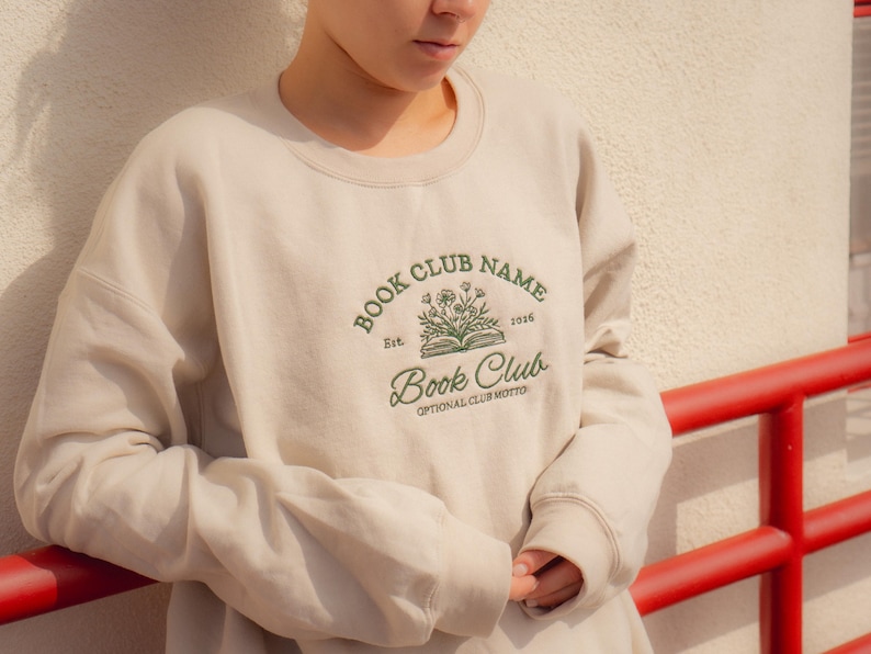 May include: A beige crewneck sweatshirt featuring dark green embroidery on the chest. The design includes an open book with flowers, the text "BOOK CLUB NAME", "Est. 2026", "Book Club", and "OPTIONAL CLUB MOTTO" in a mix of serif and script fonts.