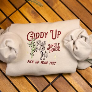May include: A beige sweatshirt with the text "Giddy Up Jingle Horse Pick Up Your Feet" embroidered in red on the front.